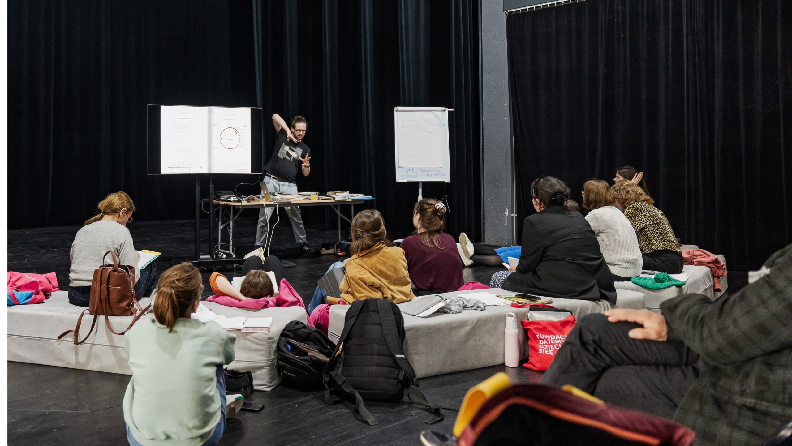 Osoby na scenie teatralnej siedzące na pufach i na podłodze, słuchające mężczyzny prowadzącego warsztaty. 
