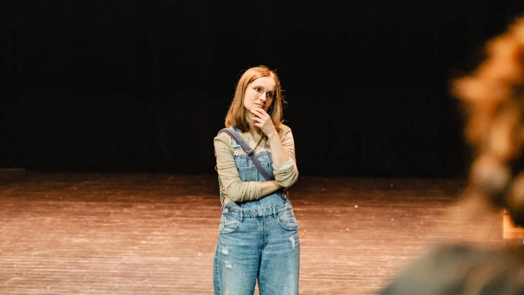 Kobieta w ogrodniczkach stoi na scenie teatralnej.